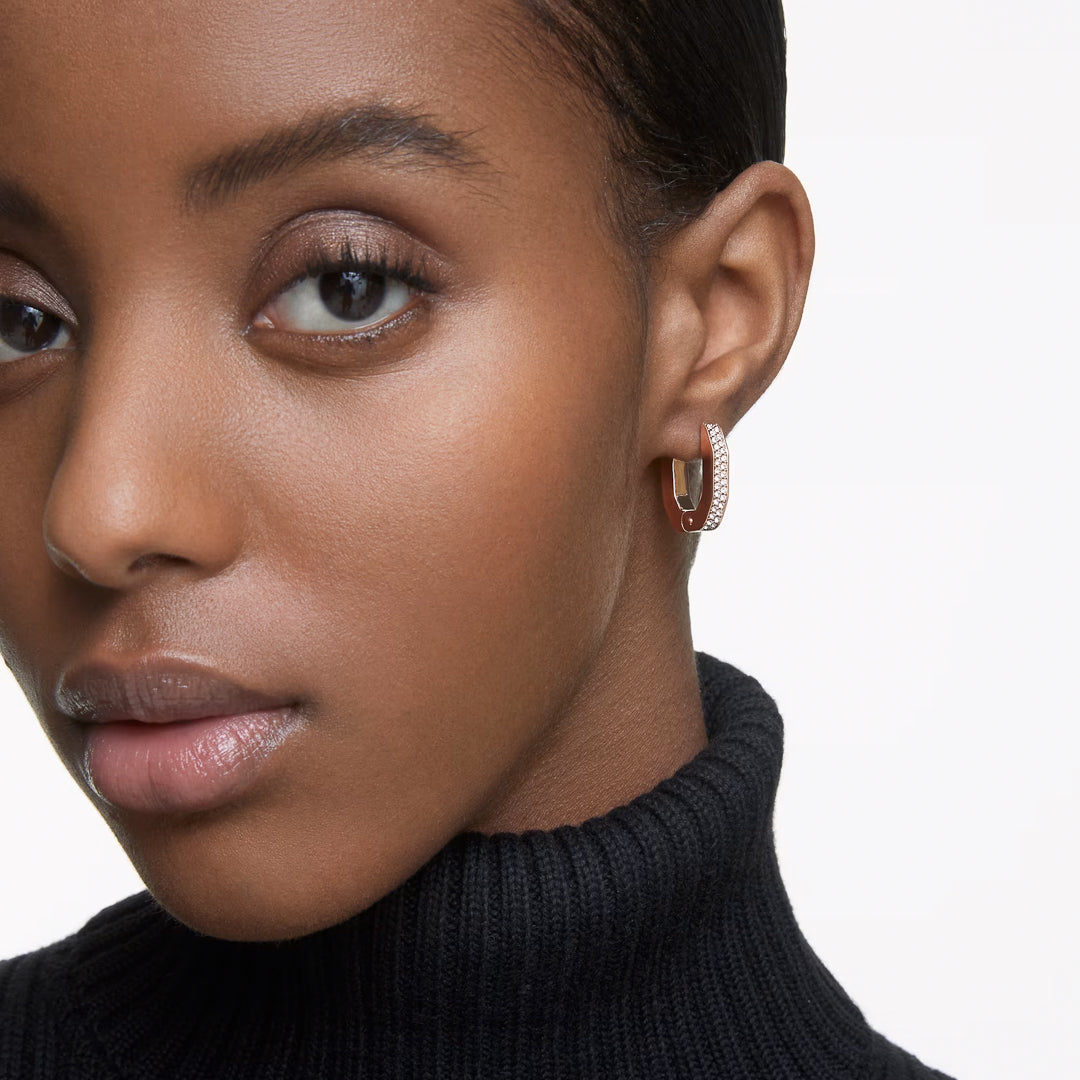 A close-up of a person wearing a black turtleneck and Swarovski Dextera Hoop Earrings in rose gold-tone. The plain white background highlights their smooth skin and subtle makeup.