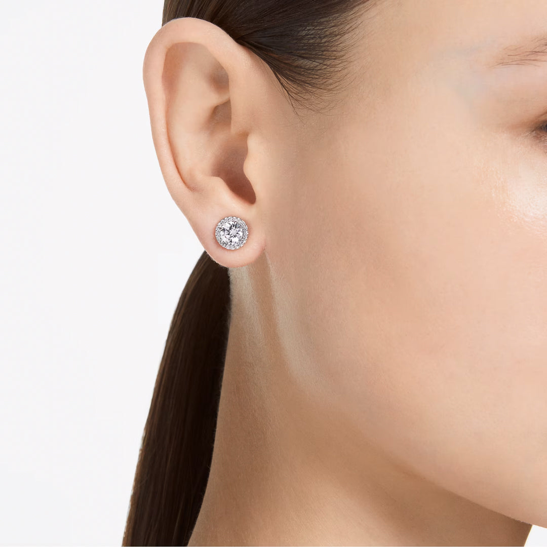 Close-up of a person's ear adorned with a pair of Swarovski Una Angelic Stud Earrings. The person's hair is pulled back, highlighting the shimmering rhodium-plated earrings. The background is neutral, emphasizing the elegant jewelry by Swarovski.