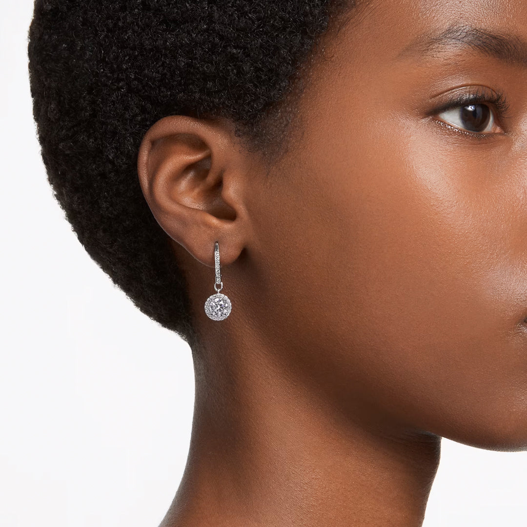 Close-up of a person with short hair wearing an elegant pair of Swarovski Una Angelic Drop Earrings. The rhodium-plated earrings, adorned with sparkling crystals, hang gracefully from small hoops against a plain white background.