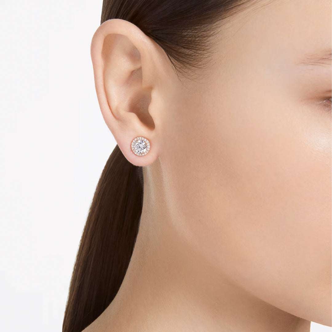 A close-up of a woman's ear showcases the Swarovski Una Angelic Stud Earrings, featuring a round diamond in a rose gold-tone setting. Her hair is pulled back, accentuating the simplicity and elegance of this exquisite piece against her skin. The plain white background ensures that all attention is directed towards these stunning earrings from Swarovski.