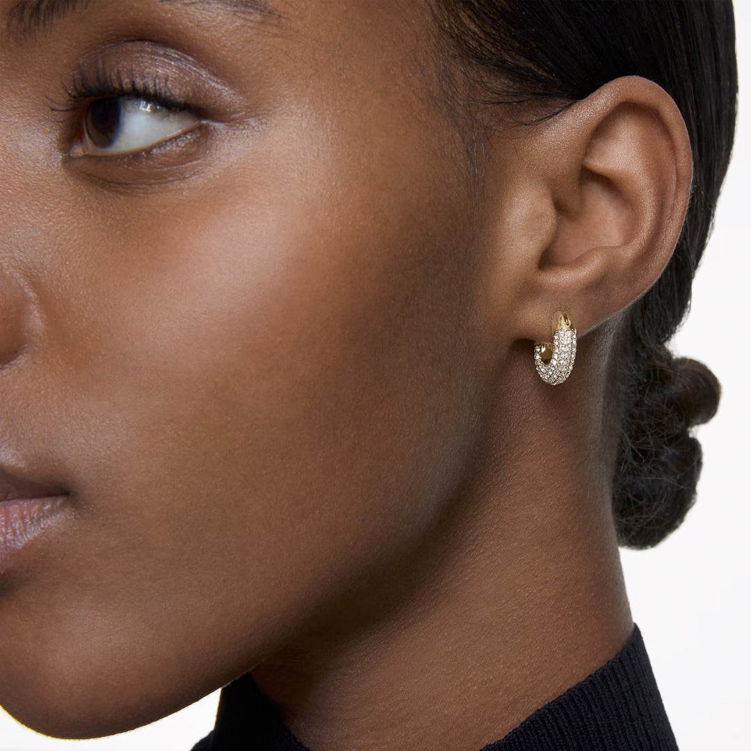 Close-up of a person wearing Swarovski Dextera Hoop Earrings. The gold-toned earrings are embellished with small gemstones, reminiscent of Swarovski Pavé. The person has smooth skin and is dressed in a black top, with their hair pulled back in a sleek bun.