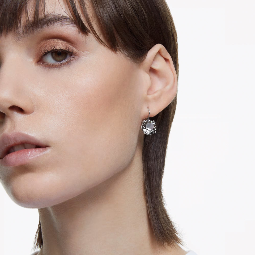 Close-up of a person with short brown hair wearing the Swarovski Millenia Drop Earrings, which are beautifully crafted from round-cut crystals. The earrings stand out against their light skin and minimal makeup, featuring neutral eyeshadow and light pink lipstick. The background is plain white.