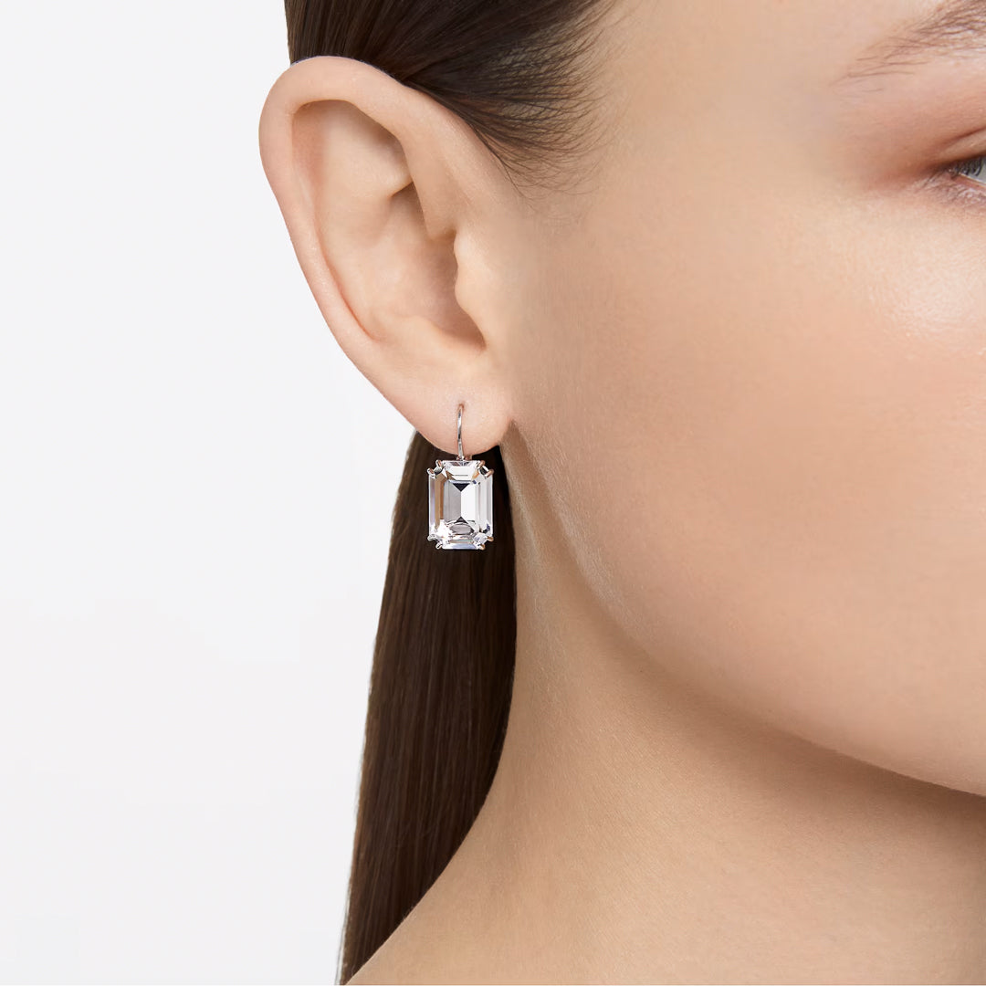 A close-up captures someone wearing the dazzling Swarovski Millenia Drop Earrings from the renowned brand Swarovski. Adorned with octagon cut crystals, this rhodium-plated piece stands out beautifully against their straight hair and skin, while a plain white background accentuates the exquisite jewelry.
