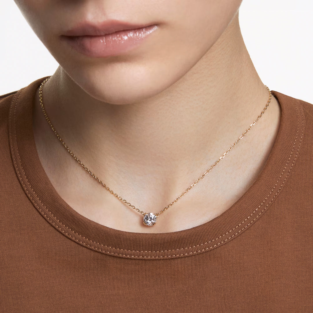 A person wearing a brown shirt is shown from the neck down, showcasing the Swarovski Stilla Necklace Pendant by Swarovski, which features a gold-tone plated chain with a small, sparkling zirconia pendant.