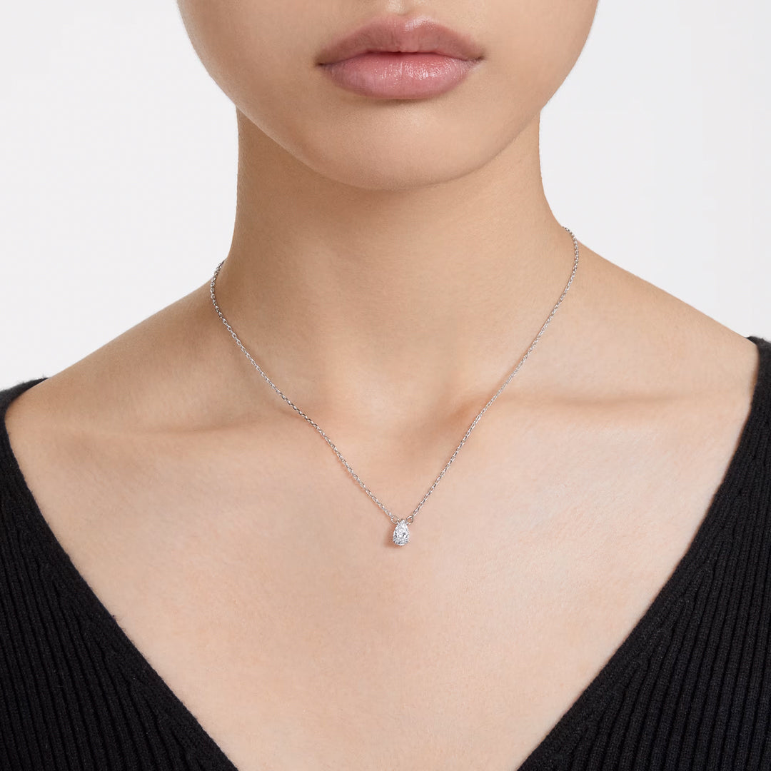 A person is adorned with a Swarovski Stilla Necklace Pendant featuring a delicate design, crafted by the renowned brand, Swarovski. Dressed in a black, deep V-neck top, the focus remains on the rhodium-plated necklace and pendant, their beauty enhanced against the softly blurred background.