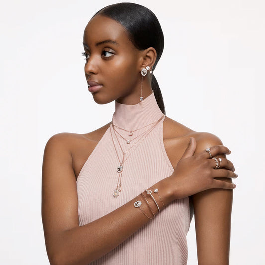 A woman wearing a sleeveless, high-neck beige top layers her necklaces, including the Swarovski Stilla Necklace Pendant featuring rose gold-tone plating and octagon-cut stones. Her look is completed with earrings, bracelets, and rings embellished with crystals as she gazes to the side against a plain white background.