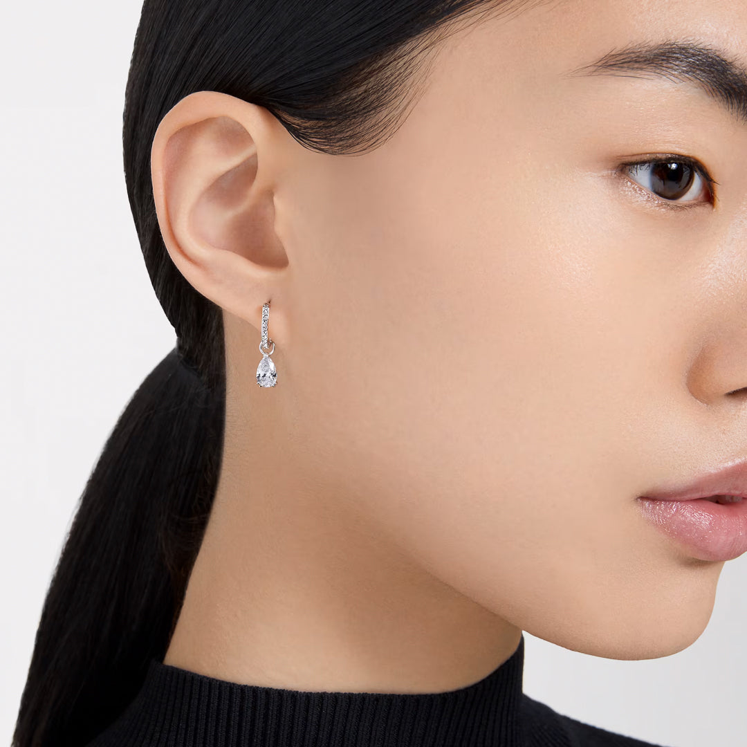 A person with long dark hair pulled back is wearing a Swarovski Stilla Stud Earring on their ear. They are dressed in a high-neck black top, with the earring's rhodium plating from Swarovski glinting against the plain white background.