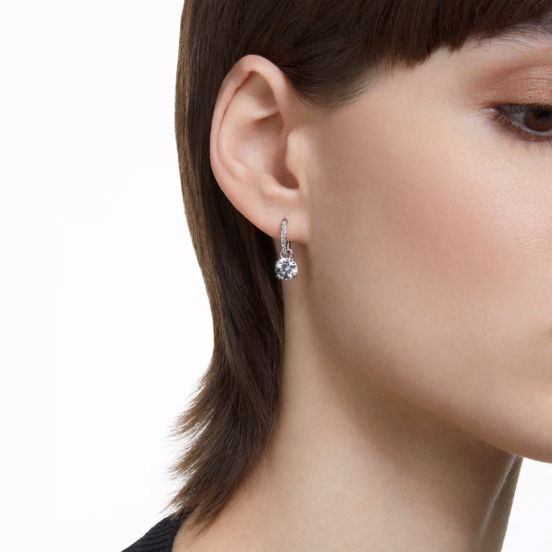 A close-up features a person with short brown hair wearing a Swarovski Stilla Drop Earring, elegantly accented with rhodium plating, on their left ear. The plain white background highlights the side profile of the person's face and ear.