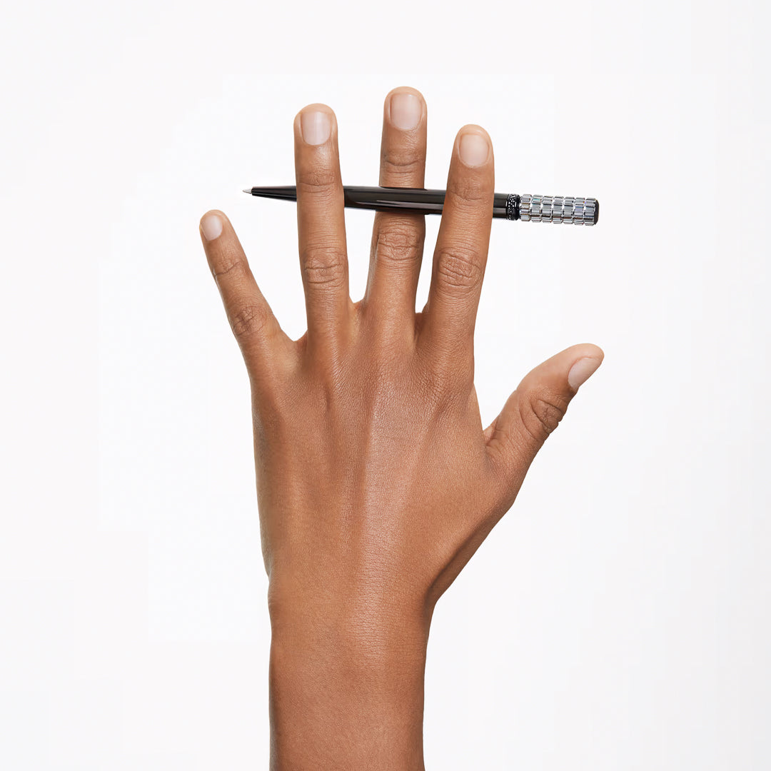A hand holds a Swarovski Ballpoint Pen, branded by Swarovski, stylishly balanced between the ring and middle fingers against a plain white background. The fingers are slightly spread, and the pen is horizontal, creating a minimalistic and creative composition.