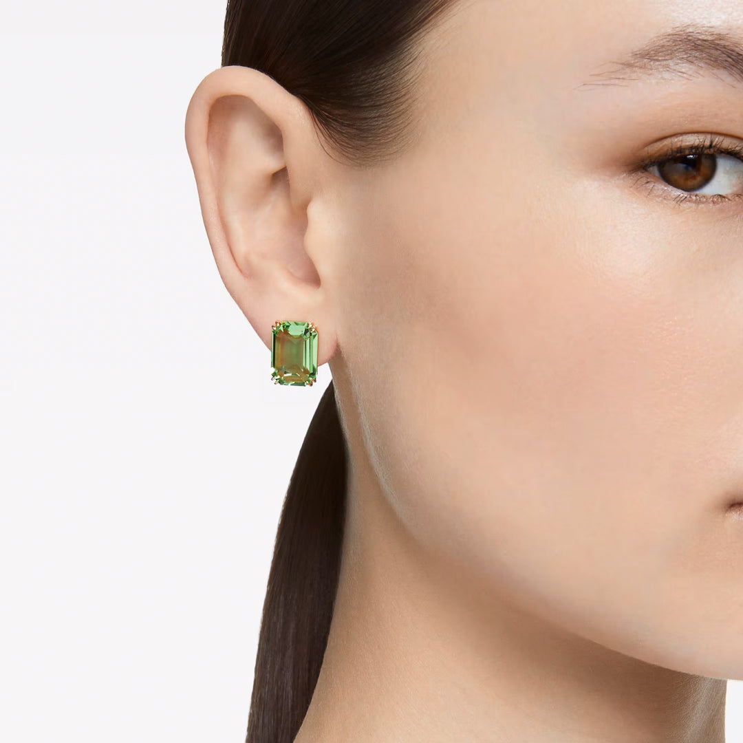 A close-up image captures a person's ear wearing the Swarovski Millenia Stud Earrings, featuring a rectangular green crystal in a prong setting. The individual's smooth skin and neatly tucked straight dark hair are set against a plain white background.