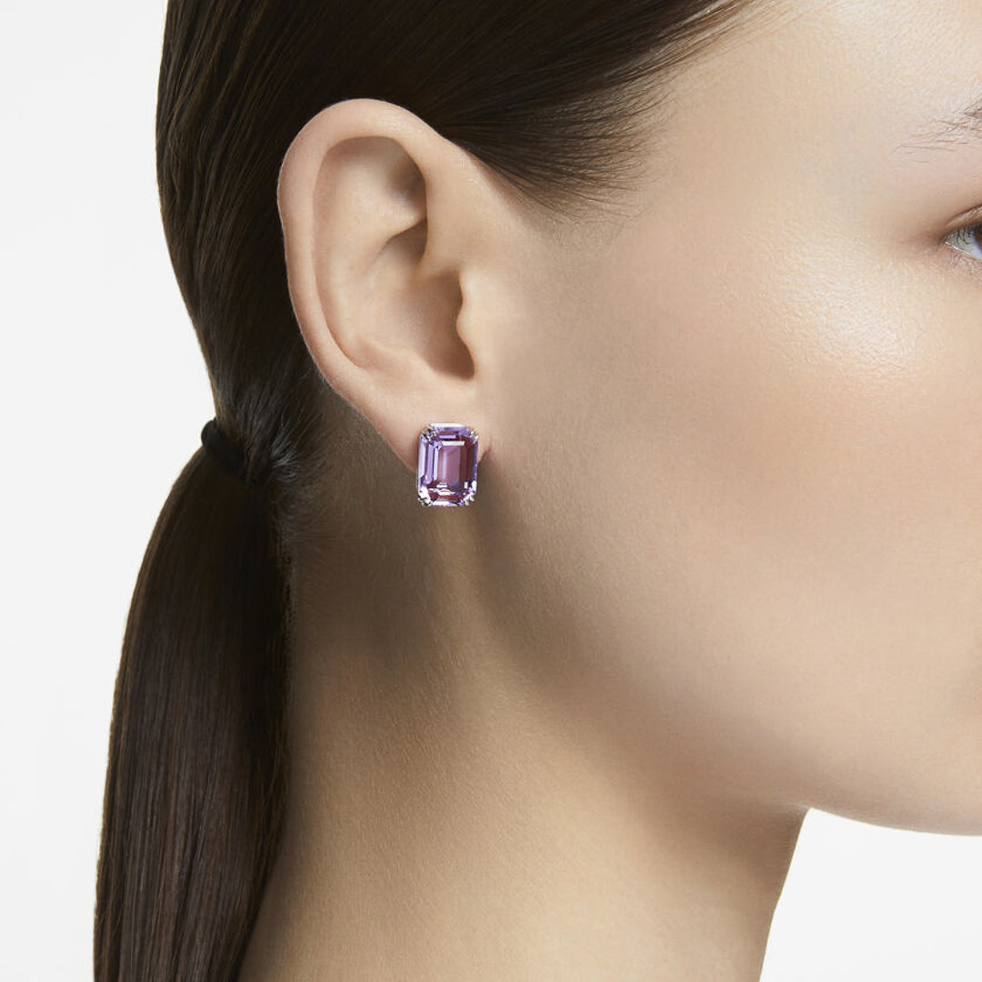 A person with dark hair pulled back is wearing the striking Swarovski Millenia Stud Earrings, featuring a large violet crystal. The rhodium-plated setting by Swarovski enhances its elegance against the plain white background.