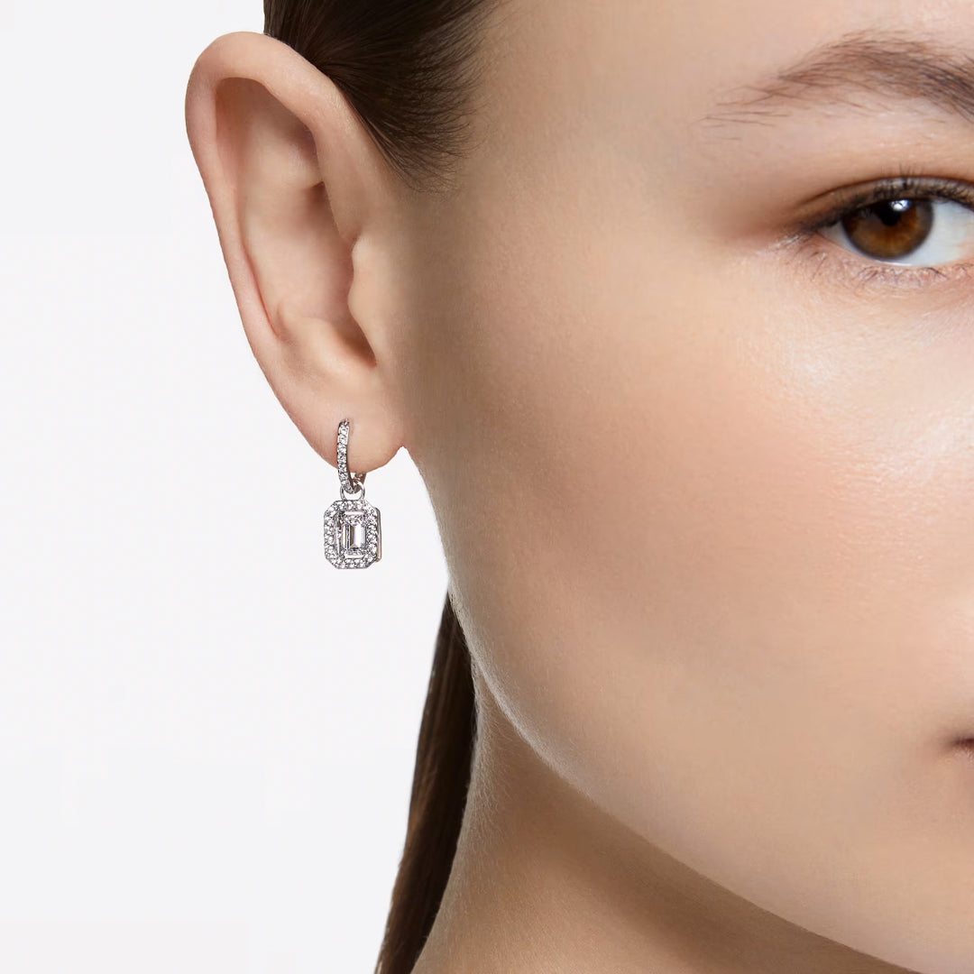 Close-up of a person wearing Swarovski Una Drop Earrings. The earrings, featuring octagon-cut stones in a rectangular design, are set in a classic style. The rhodium-plated finish gleams as the person's ear and part of the cheek are visible against a neutral background.