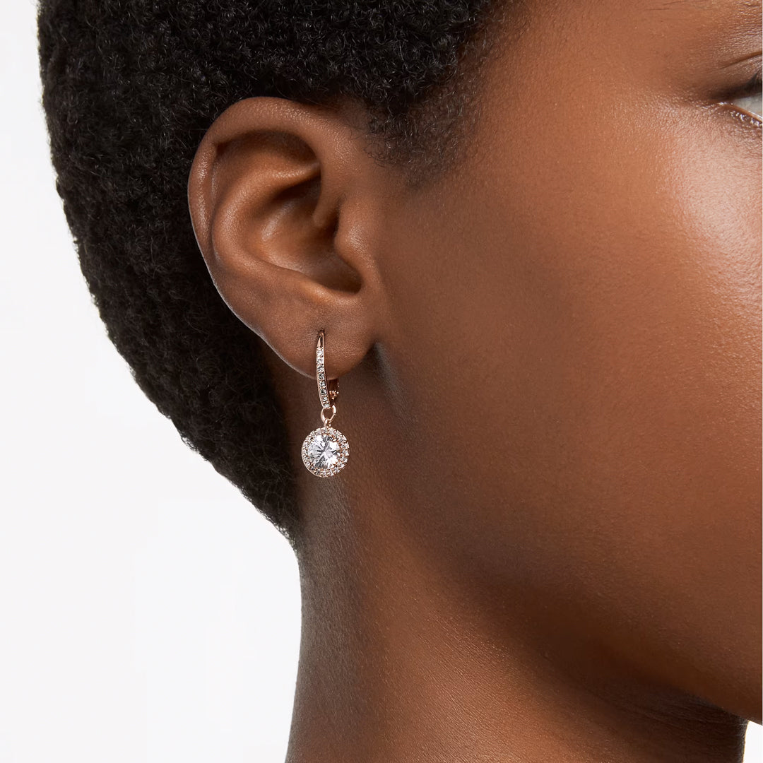 A close-up of an ear showcases the Swarovski Una Angelic Drop Earrings, which feature a large round gemstone in a delicate rose gold-tone setting. These Swarovski earrings, embedded with crystals, hang gracefully from a small hoop, while the person's short hair accentuates the jewelry's brilliance.