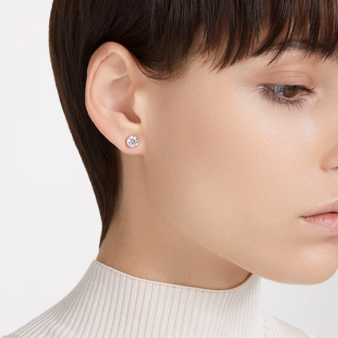 Close-up of a person with short dark hair, wearing a white turtleneck sweater and an elegant Swarovski Stilla Stud Earring in their left ear. The background is plain, drawing focus to the sparkling earring and refined facial profile.