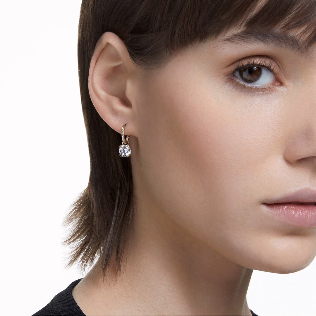 Close-up of a person with short brown hair wearing a large Swarovski Stilla Drop Earring. The image captures the right side of the face against a white background, highlighting the earring’s sparkle and gold-tone plated finish.