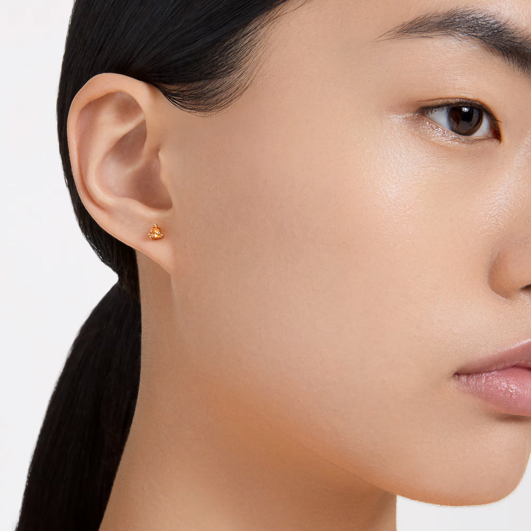 Close-up of a person with long dark hair tied back, wearing Swarovski Chroma Stud Earrings by Swarovski. These small gold stud earrings are adorned with trilliant-cut orange crystals. The plain white background highlights the smooth complexion and this standout accessory.