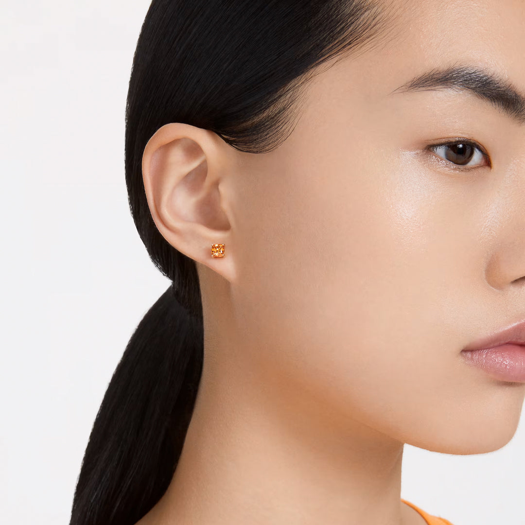 Close-up of a person wearing the Swarovski Chroma Stud Earrings, designed as small flower-shaped studs in vibrant orange and elegantly finished with gold-tone plating. Their dark hair is neatly pulled back, and they are facing slightly to the left against a simple backdrop.