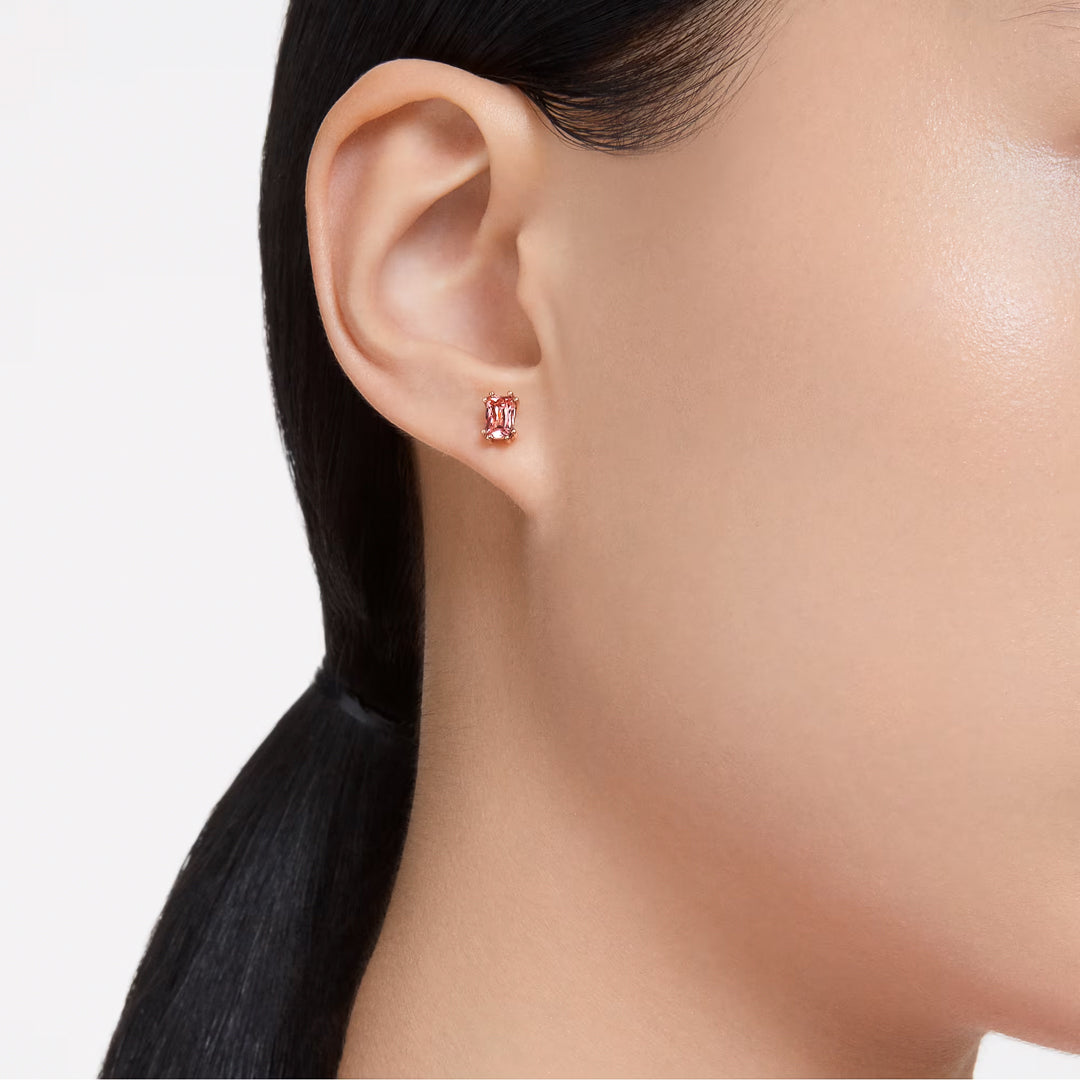 Close-up of a person's right ear adorned with Swarovski Chroma Stud Earring, featuring a small, rectangular pink zirconia in a rose gold-tone setting. The person has dark hair pulled back into a sleek style against a plain white background.