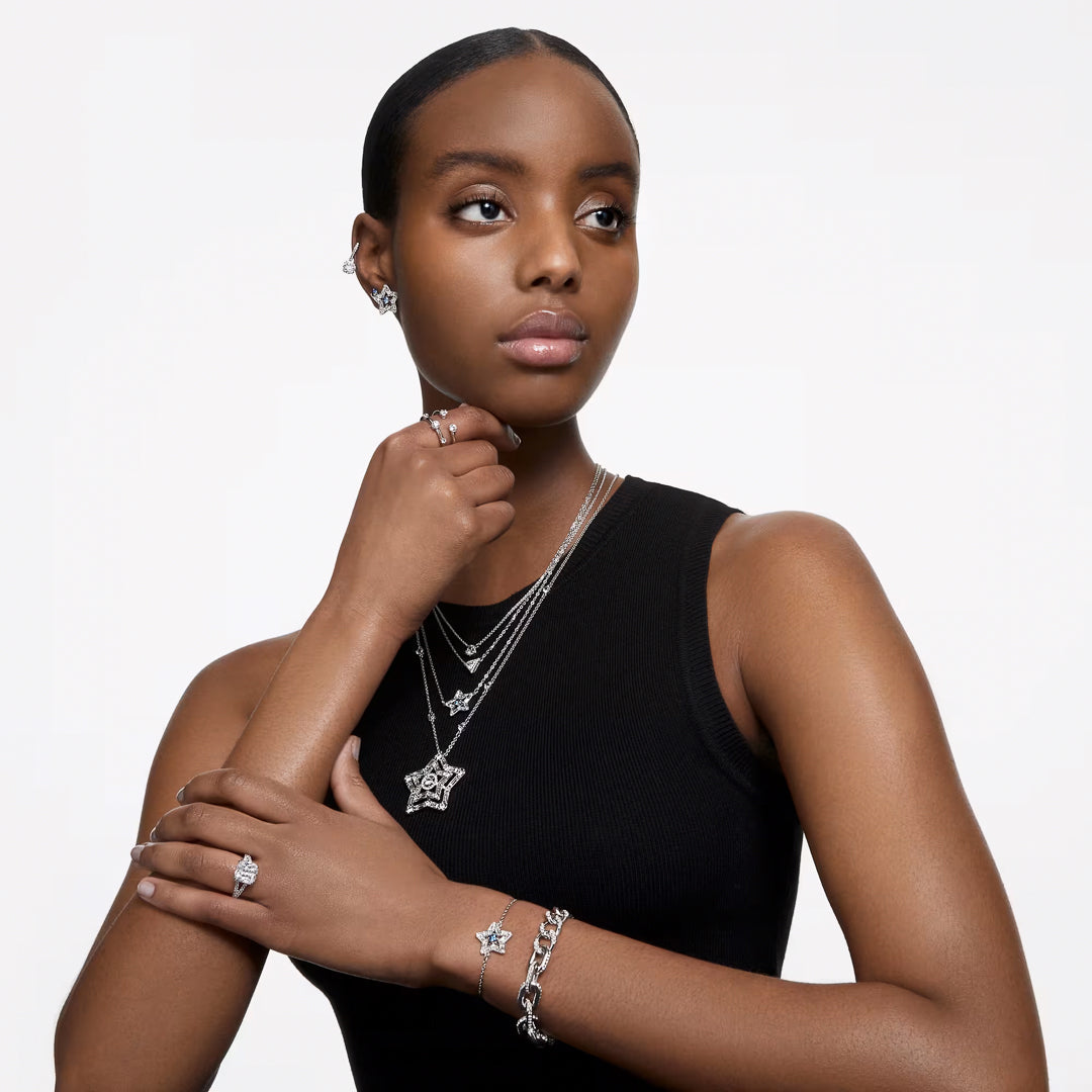 A woman wearing a black sleeveless top adorned with elegant Swarovski Stilla Stud Earrings and other rhodium-plated silver jewelry, including a star-shaped pendant necklace, gazes thoughtfully to the side, her hand gently placed under her chin.