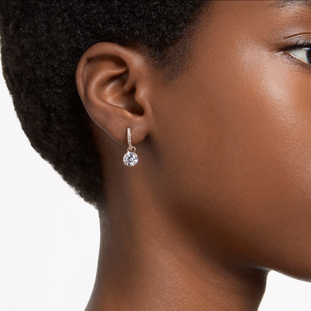 Close-up of a person wearing the Swarovski Stilla Drop Earrings by Swarovski, showcasing a sparkling teardrop design with rose gold-tone plating and a small hoop. These earrings feature a large, clear gemstone adorned with intricate crystals. The person has short, curly hair and smooth skin set against a plain white background.