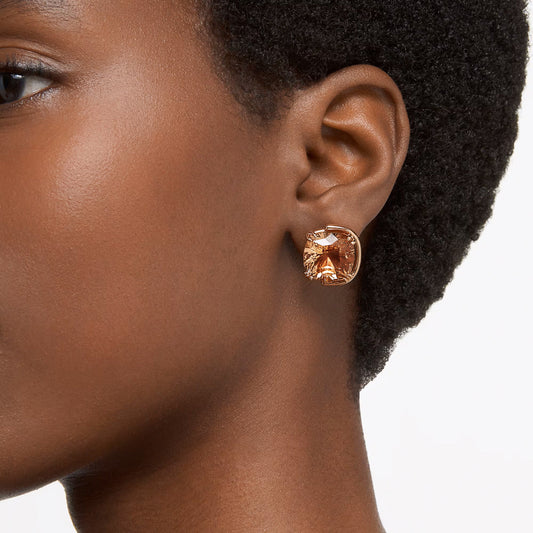 Close-up of a person wearing the Swarovski Harmonia Stud Earrings, which feature an amber-colored stone cut in a geometric shape and plated in gold-tone. The earring stands out against their dark hair and skin, capturing the elegance characteristic of Swarovski creations.