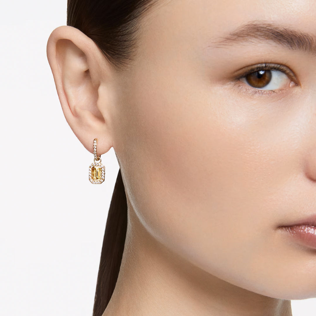 A close-up shot features an individual wearing Swarovski Una Drop Earrings, which are rectangular in shape and elegantly dangle with a gold setting and pavé crystals. The white background highlights the jewelry, while the person's long brown hair is tied back to reveal a clear side profile, showcasing the elegance of their pierced earrings from Swarovski.