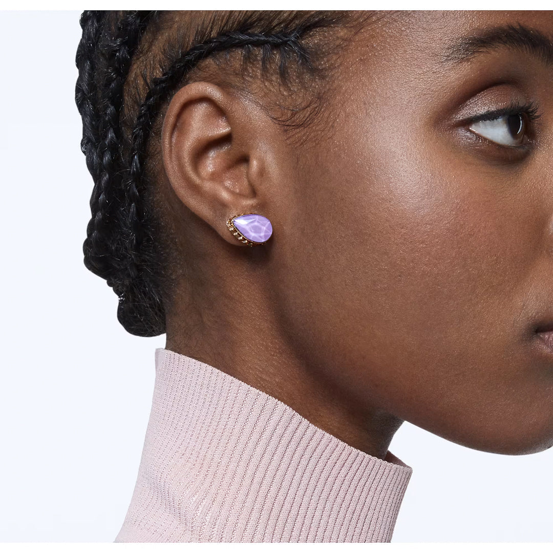 A person's profile is depicted wearing stunning lavender Swarovski Chroma Stud Earrings. They sport braided hair and are clad in a light pink turtleneck sweater, set against a plain white background that beautifully highlights the elegant drop-cut crystals.