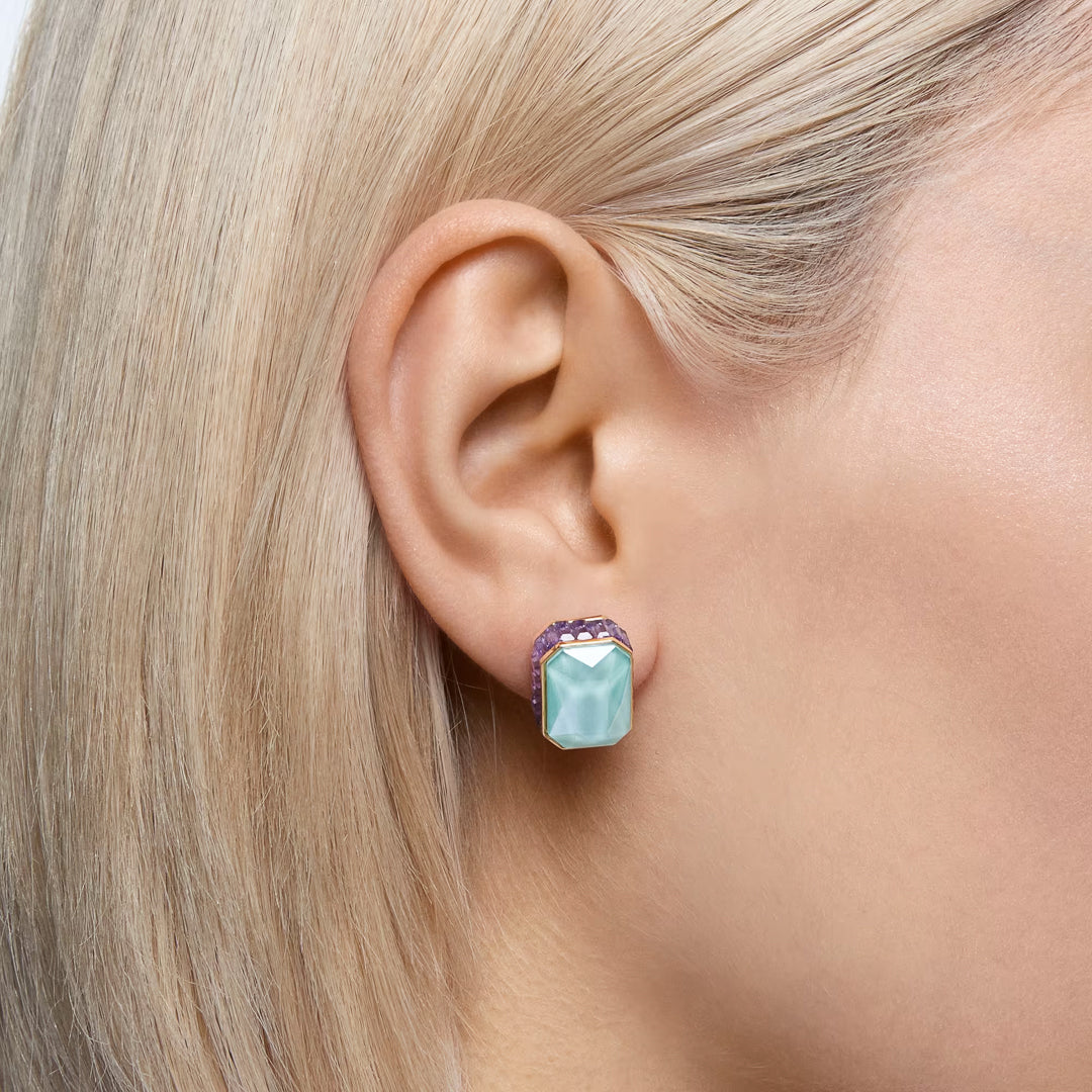 Close-up of an individual with blonde hair wearing the exquisite Swarovski Chroma Stud Earrings. These earrings showcase a large, rectangular blue gemstone set in gold-tone plated metal, enhanced by pink accents. The intricate design is elegantly displayed against the ear.