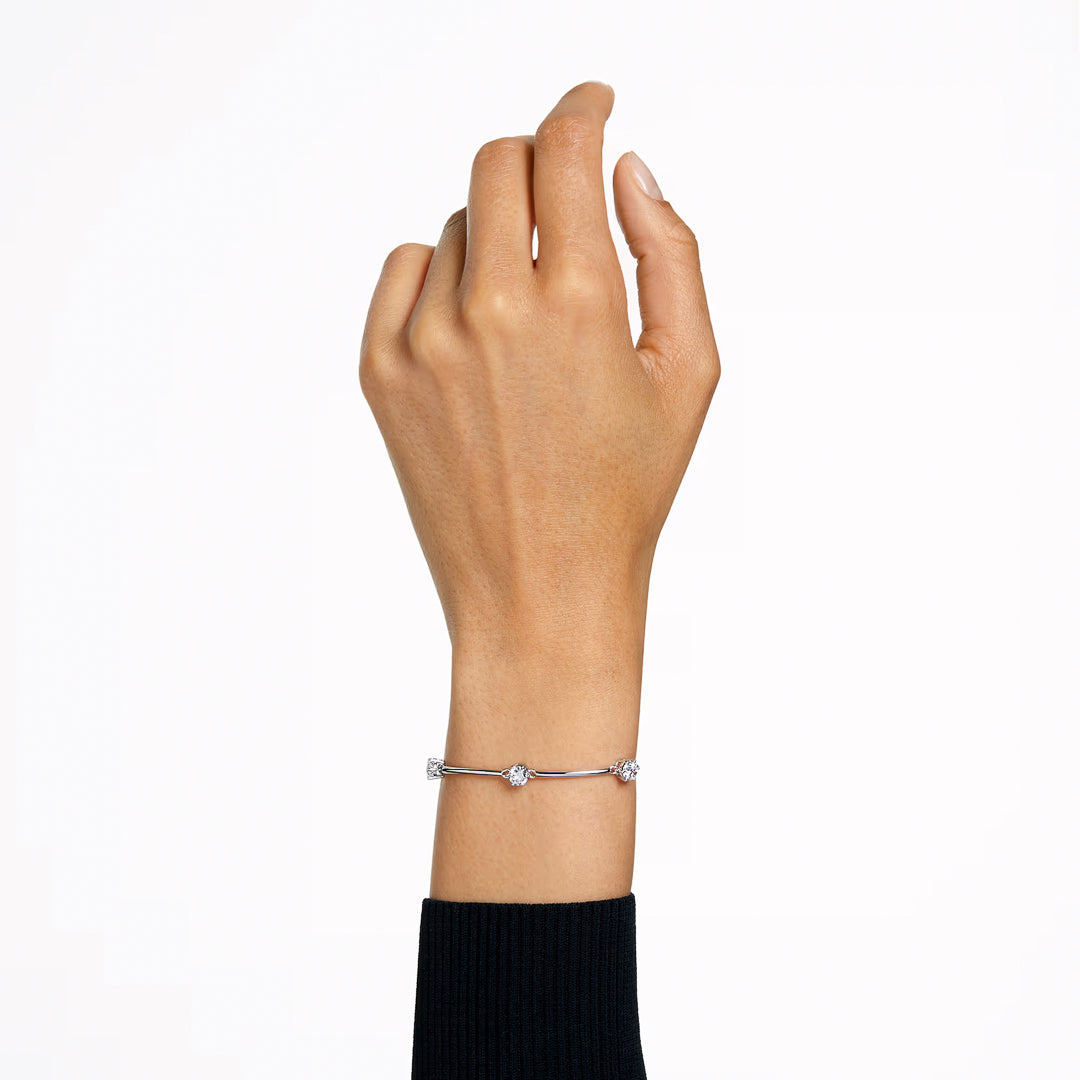 A hand wearing the sophisticated Swarovski Constella Bangle, a delicate silver bracelet with small round charms and zirconia accents, is raised against a plain white background. The rhodium-plated design complements the long-sleeve black sweater, enhancing the elegant appearance.