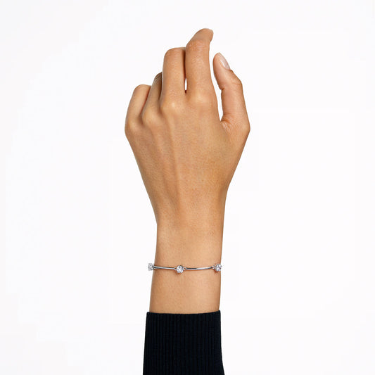 A hand wearing the sophisticated Swarovski Constella Bangle, a delicate silver bracelet with small round charms and zirconia accents, is raised against a plain white background. The rhodium-plated design complements the long-sleeve black sweater, enhancing the elegant appearance.