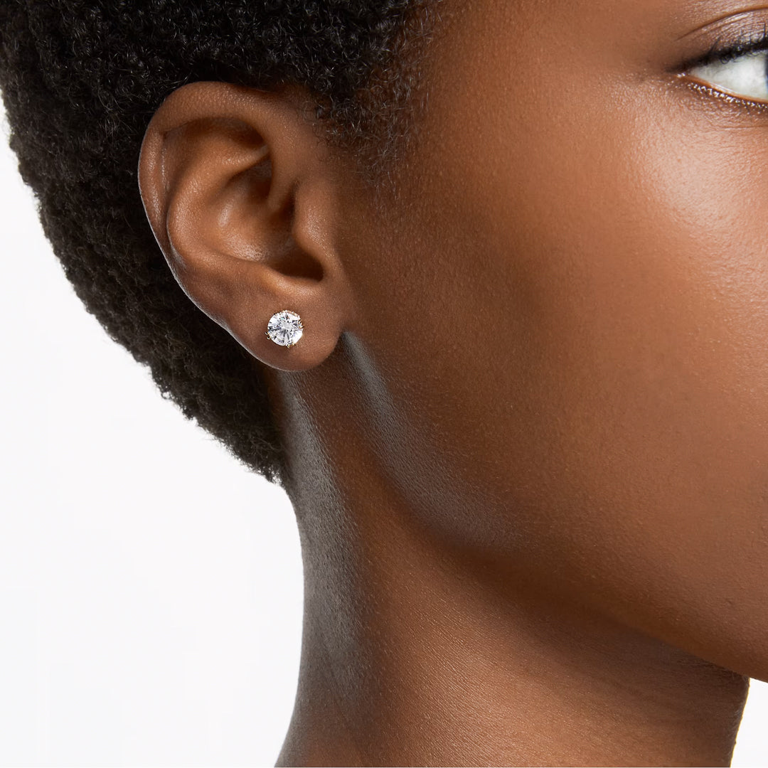 A close-up of a person's ear adorned with a Swarovski Stilla Stud Earring, featuring sparkling zirconia against a plain background. The gold-tone plating from Swarovski adds elegance, while the person's short hair and smooth skin beautifully complement the look.