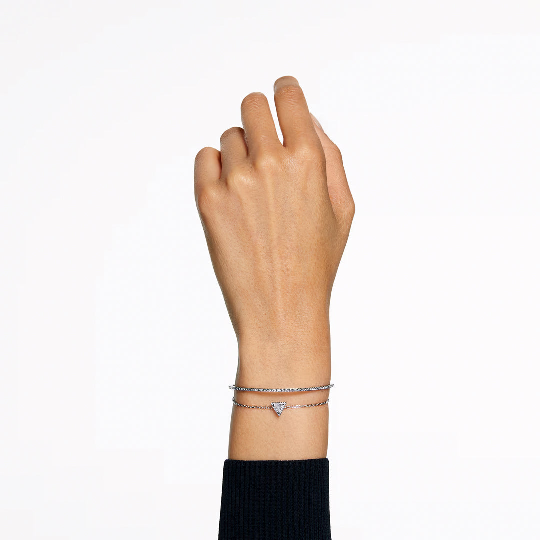 A left hand is shown against a white background, elegantly accessorized with two delicate silver bracelets. One bracelet is the Swarovski Stilla Bracelet, characterized by its small triangular charm, while the person is dressed in a black long-sleeve top.