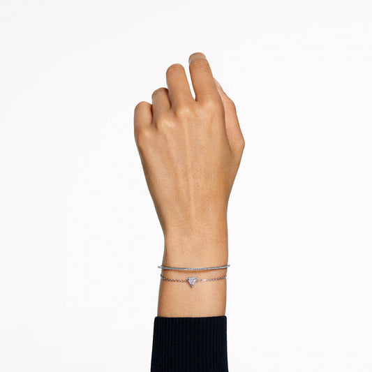 A left hand is shown against a white background, elegantly accessorized with two delicate silver bracelets. One bracelet is the Swarovski Stilla Bracelet, characterized by its small triangular charm, while the person is dressed in a black long-sleeve top.