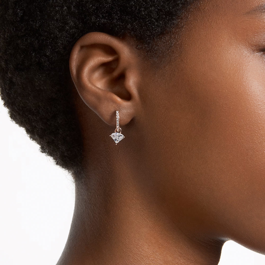 Close-up of a person wearing Swarovski Stilla Drop Earrings in rose gold-tone, featuring triangle-cut crystals. A small, sparkling diamond dangles gracefully from a simple, thin hook. The plain white background enhances the elegance of these Swarovski earrings and complements the person's ear.