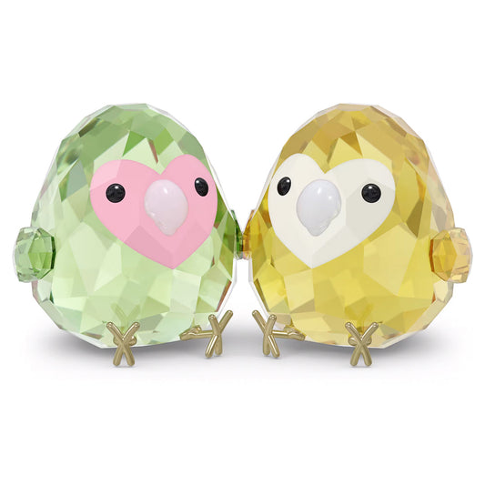 The Swarovski All you Need are Birds Love Bird Couple Home Décor features two crystal figurines with large eyes and metallic feet, positioned side by side. One is adorned in vibrant green and pink hues, while the other sparkles in yellow and white. Their faceted, gem-like appearance brilliantly catches the light, capturing the enchanting charm of colorful birds.