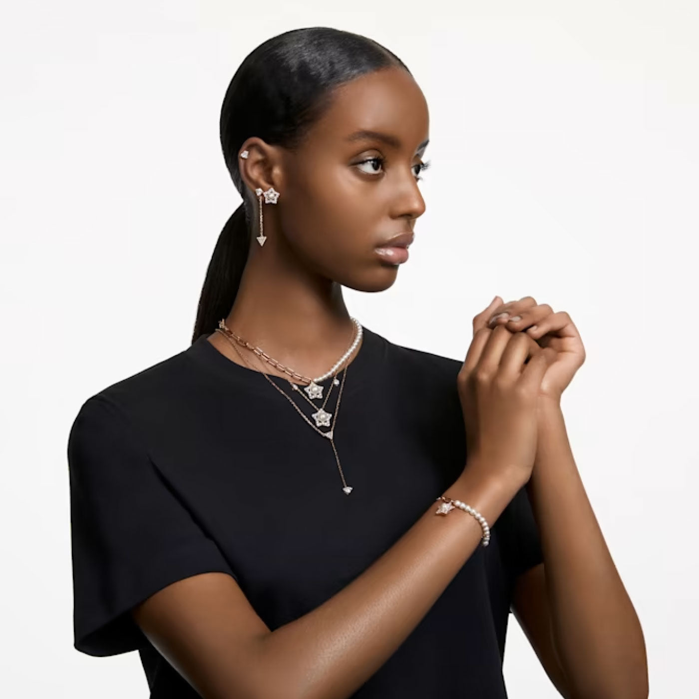 A woman with a sleek hairstyle poses against a plain background. She wears a black top and elegant jewelry, featuring earrings with a star motif, layered necklaces, and the Swarovski Stella Bracelet adorned with crystal pearls. Her hands are gently clasped together.