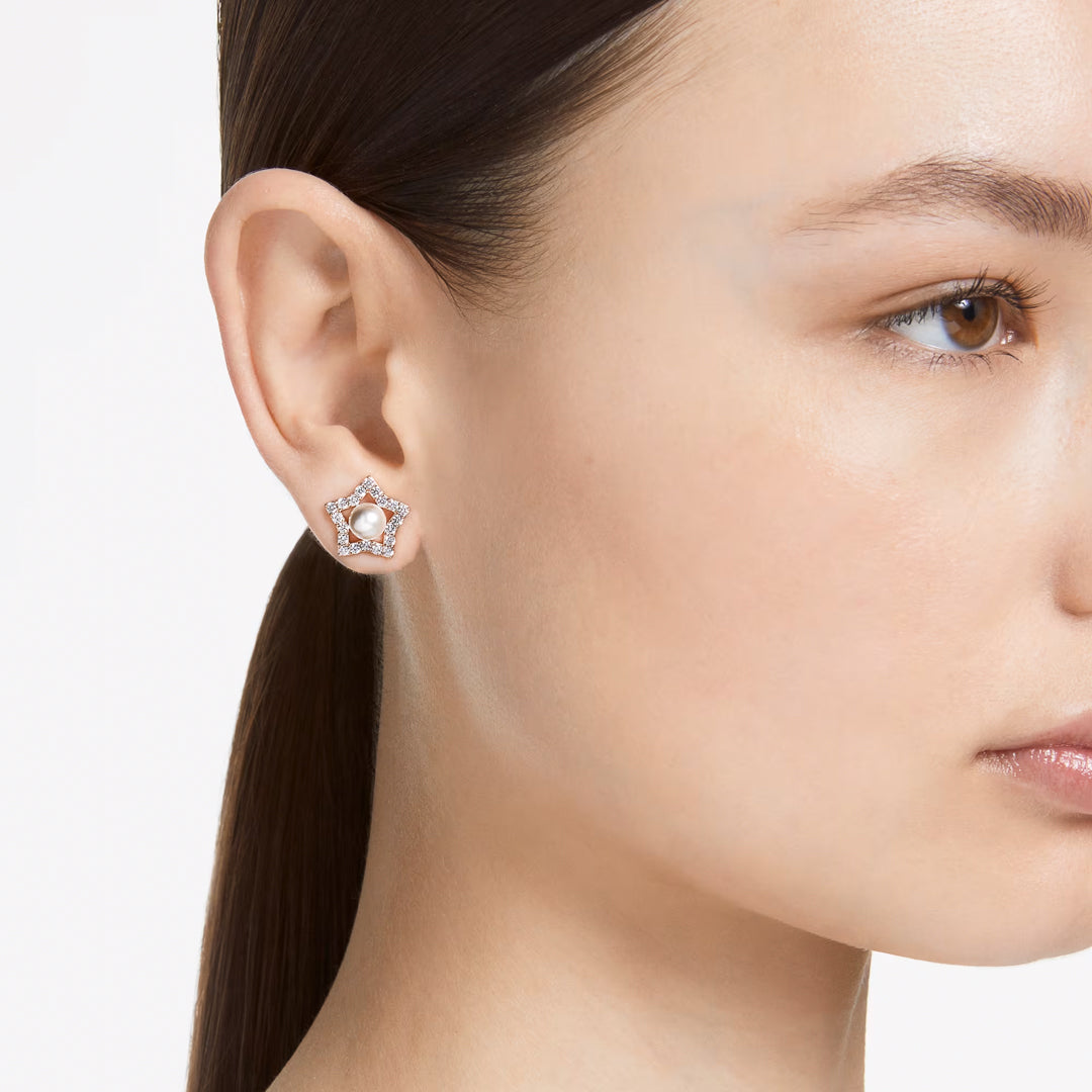 A close-up shows an individual wearing the Swarovski Stella Stud Earrings, featuring a star-shaped design with rose gold-tone plating. These earrings sparkle subtly against their ear, framed by dark hair pulled back to reveal smooth, clear skin. The plain white background accentuates the elegance of the Swarovski piece.
