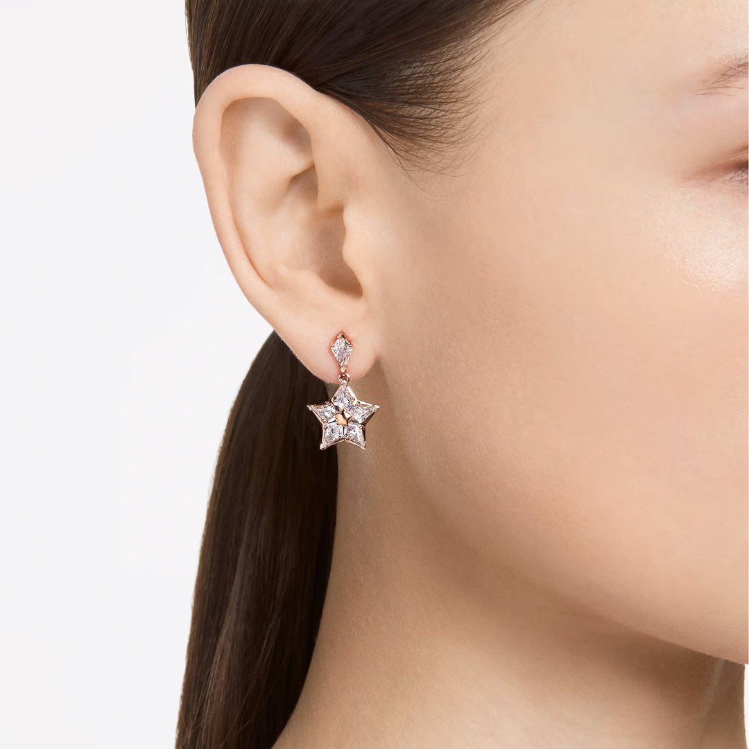 A close-up image showcases a person's ear wearing the Swarovski Stella Drop Earrings by Swarovski, featuring star-shaped, rose gold-tone design adorned with shimmering gemstones. The jewelry has a shiny, metallic appearance and is attached to a small round Zirconia stone, set against a plain white background.