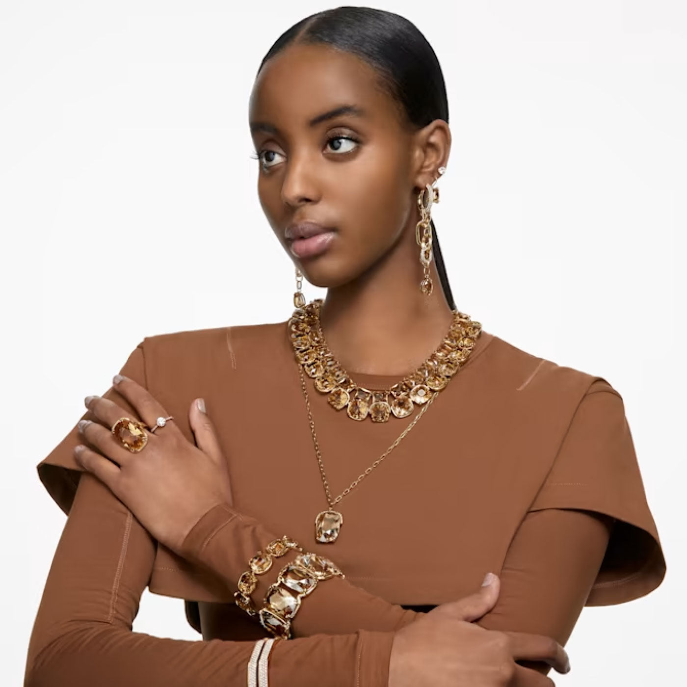 The individual, who has long, dark hair styled back, is dressed in a brown outfit and adorned with multiple pieces of gold jewelry. Their accessories include the Swarovski Harmonia Necklace Pendant from the Swarovski brand, along with earrings, rings, and bracelets. They stand against a neutral background while looking to the side with their arms crossed.