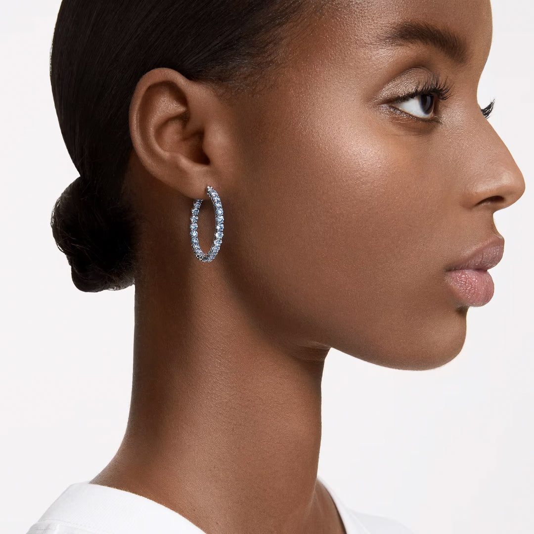 Profile of a person with smooth skin and tied-back hair, showcasing striking Swarovski Matrix Hoop Earrings adorned with blue stones. The backdrop is white, and they are wearing a white top.