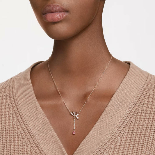 A person wearing a beige knit sweater is shown from the shoulders up, displaying a romantic gift: the Swarovski Hyperbola Y Necklace Pendant, crafted by Swarovski. This delicate rose gold necklace showcases a striking bow design and features a drop pendant embellished with clear crystals. The background is plain white.