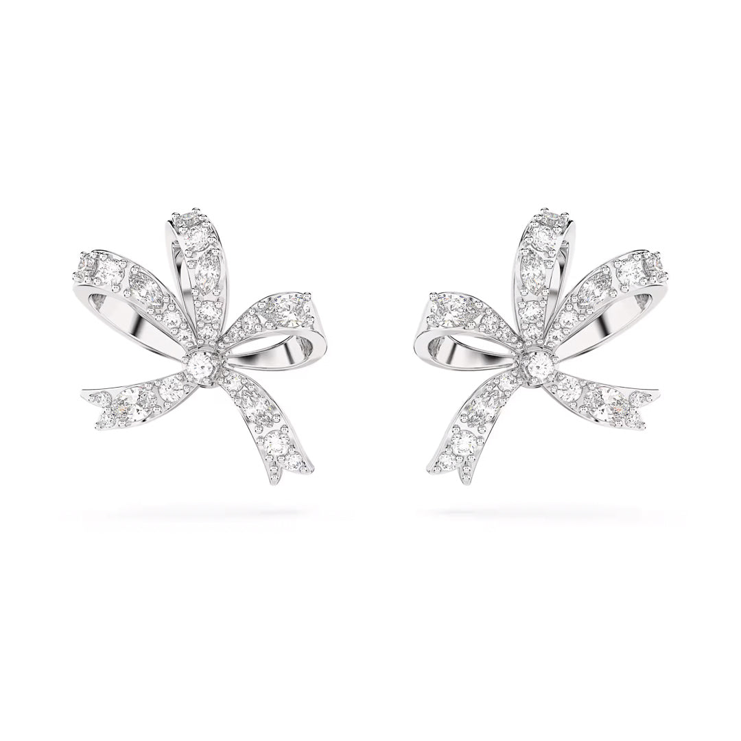 The Swarovski Hyperbola Stud Earrings by Swarovski are rhodium-plated and feature a bow-shaped design adorned with sparkling diamonds. These earrings reflect light beautifully, creating a shimmering effect, and include intricate ribbon-like loops and tails that highlight elegance and sophistication.