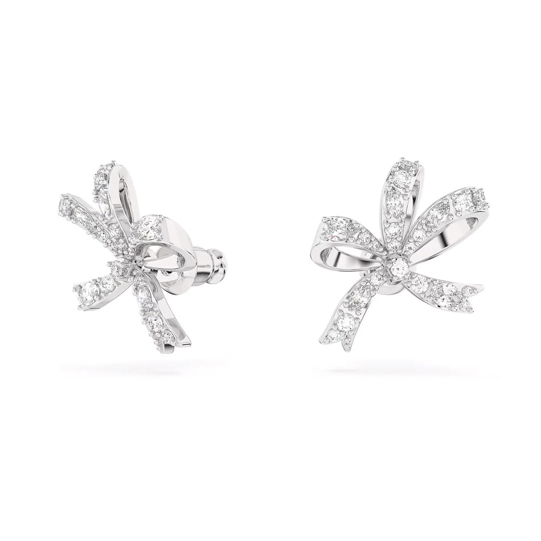 The Swarovski Hyperbola Stud Earrings by Swarovski are a pair of rhodium-plated, silver bow-inspired studs embellished with small sparkling diamonds and clear crystals. They boast intricate detailing and a glossy finish, with the symmetrical design elegantly set against a plain white background.