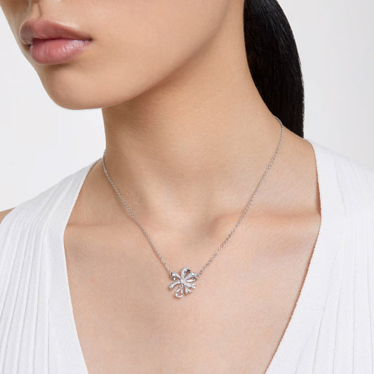 A person is shown from the nose to the shoulders wearing a white top, showcasing the Swarovski Hyperbola Necklace Pendant, which is rhodium-plated and adorned with clear crystals in a decorative bow shape. The hair is styled pulled back.