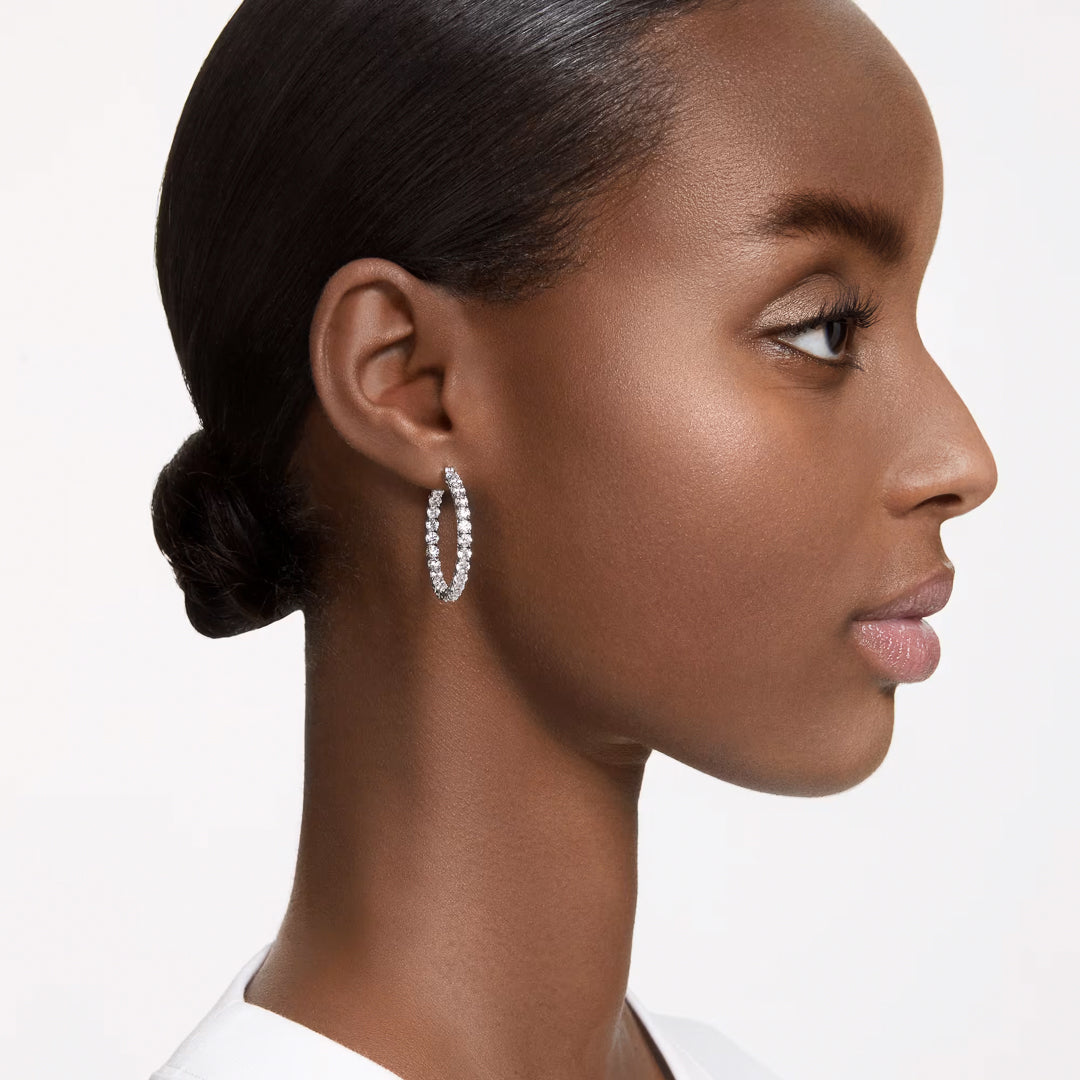 A woman with smooth, dark hair styled in a low bun is wearing the stunning Swarovski Matrix hoop earrings. The rhodium-plated design with zirconia accents adds a touch of sparkle as she looks to the right with a neutral expression against a plain white background.