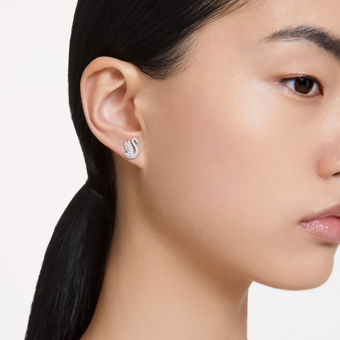Against a plain backdrop, a person with long hair has their tresses tied back, highlighting the exquisite design of a Swarovski Swan Stud Earring adorned with pavé crystals on their left ear.