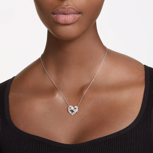 A person wearing a black top and a Swarovski Matrix Necklace Pendant, designed by Swarovski, which is silver, rhodium-plated in a heart shape with a clear gemstone. The plain white background accentuates the jewelry and the person's neckline.