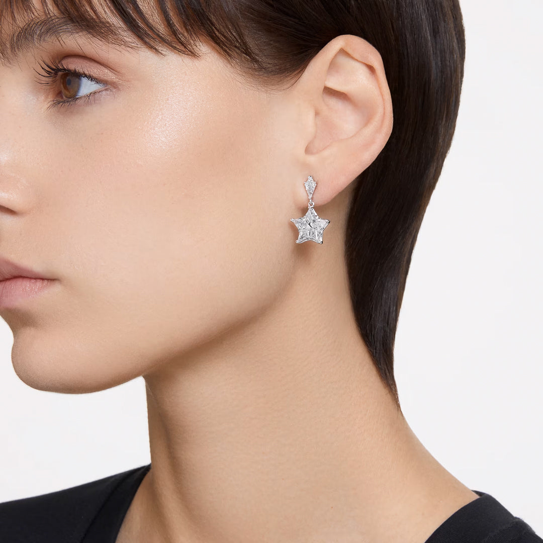 Close-up of a person with short dark hair wearing Swarovski Stella Drop Earrings, shimmering with small crystals under their rhodium plating. The individual dons a black top against a plain white background, highlighting the magical allure of the accessory by Swarovski.