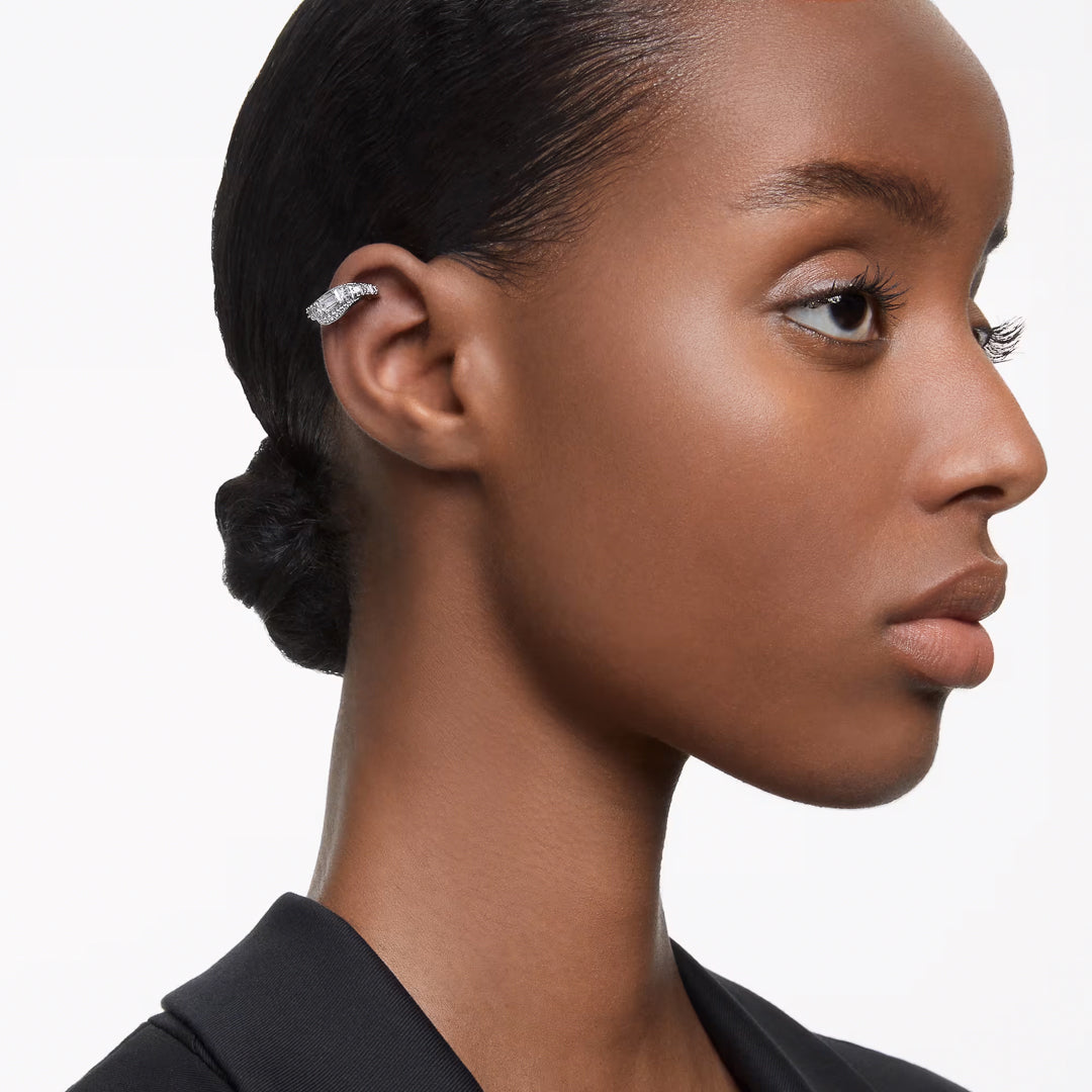 A woman with a neat bun models the Swarovski Hyperbola Clip Earrings, highlighting their unique mixed-cut design adorned with small crystals set in rhodium plating. Her expression is neutral and she is dressed in either a black top or blazer against a white background.