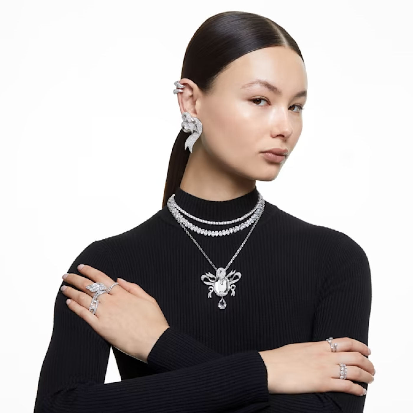 A person with dark hair tied back wears a black long-sleeve top and several silver jewelry pieces, including the Swarovski Hyperbola Cocktail Ring. Their arms are crossed, and they appear poised against a plain white background.